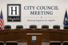 City Council meeting graphic featuring the Highfill logo on a beige background, with the headline “City Council Meeting” in large blue text and the tagline “Working Together for Highfill” beneath it. The image shows a formal council chamber with a curved wooden dais, labeled seats for mayor and council members, microphones, and U.S. and state flags on either side.