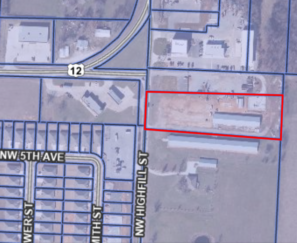 Aerial GIS map showing a section of NW Highfill Street near Highway 12. Parcel 22‑0005‑003 is highlighted with a red rectangular outline. Inside the outlined parcel is a large area of exposed, light‑colored ground and one long rectangular building along the lower right side. Surrounding parcels contain additional buildings, roads, and property boundary lines drawn in blue. Residential lots appear to the southwest, and commercial or industrial structures appear to the north.