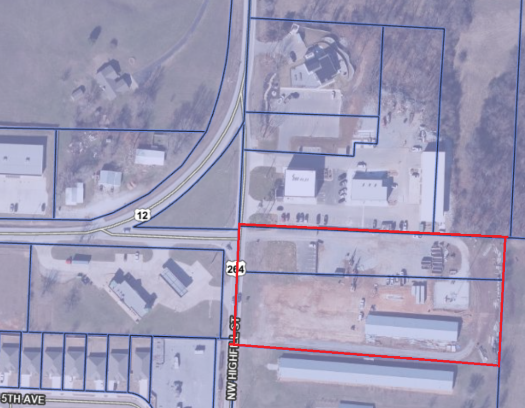 Aerial GIS map showing parcels along NW Highfill Street near Highway 12. Two adjacent parcels, 22‑00005‑003 and 22‑00004‑000, are outlined together in a single large red rectangle that spans from NW Highfill Street eastward. Within the outlined area are several visible site‑development features, including a large paved section near the street, open areas of exposed soil, construction materials, equipment, and a curved driveway or gravel loop on the eastern side. A long rectangular building sits near the lower right portion of the red‑outlined area. North of the parcels are several commercial or industrial buildings and parking areas. Highway 12 curves across the upper left of the image, and blue boundary lines outline surrounding properties. Residential lots appear to the southwest of the site.