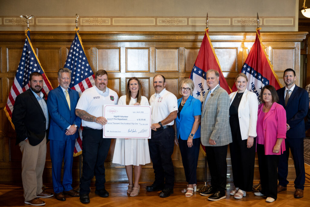 Group of people standing in a formal room at the Arkansas State Capitol in front of wooden paneling and multiple U.S. and Arkansas flags. The group is holding an oversized ceremonial check labeled ‘Highfill Volunteer Fire Department’ for a Rural Services Grant in the amount of $12,151.42. The setting appears to be the Governor’s Conference Room, and everyone is posed together for the award ceremony.